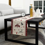 Une table basse noire avec un chemin de table décoratif représentant des rennes et des flocons de neige, accompagnée d'une horloge et d'accessoires de cuisine sur un tapis gris dans un salon cosy.