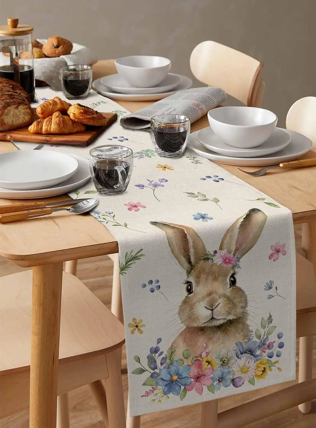 Une table dressée avec des petits pains, des tasses et une nappe décorée d'un motif de fleurs et d'un lapin.