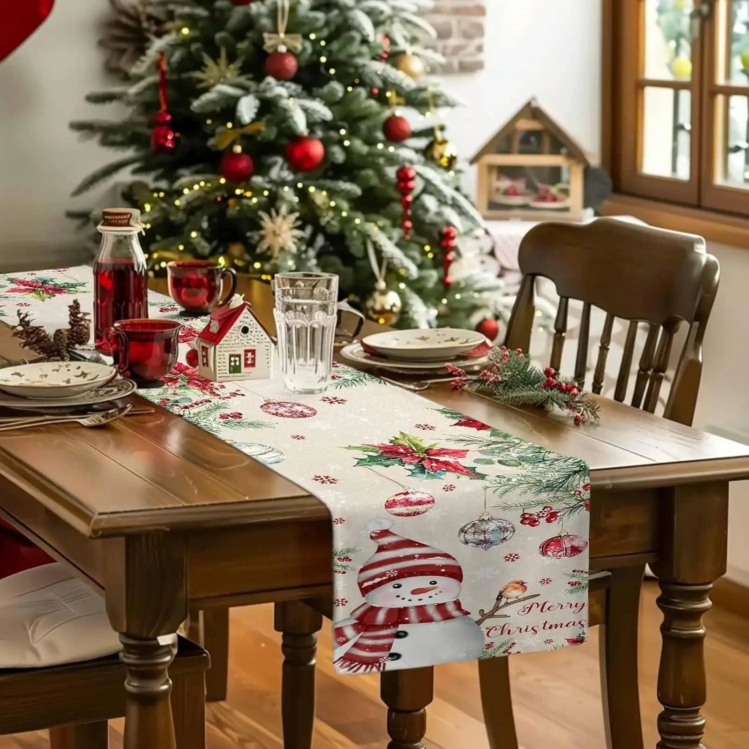 Cette image montre une table de salle à manger décorée pour Noël, avec un magnifique sapin illuminé en arrière-plan, des assiettes, des verres et un chemin de table avec des motifs de Noël.