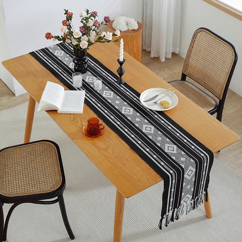 Une table en bois naturel avec une nappe noire et blanche rayée, une bougie blanche, des fleurs dans un vase et des accessoires décoratifs.