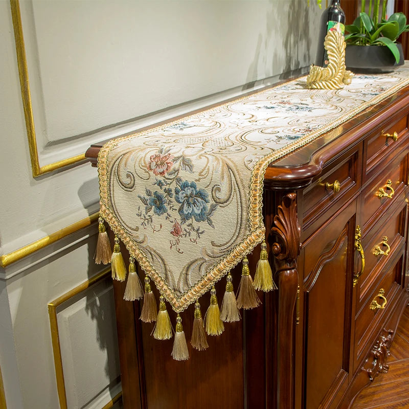 Cette image représente un meuble décoratif avec un chemin de table en tapisserie ancienne, orné de glands et de motifs floraux, posé sur une commode en bois foncé sculptée.