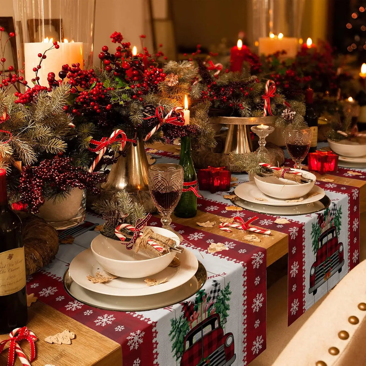 Une table de Noël joliment décorée avec des arrangements de branches de pin, de baies rouges et de bougies allumées, accompagnés de plats et de verres élégants sur un tissu rouge orné de motifs de flocons de neige et de camions vintage.
