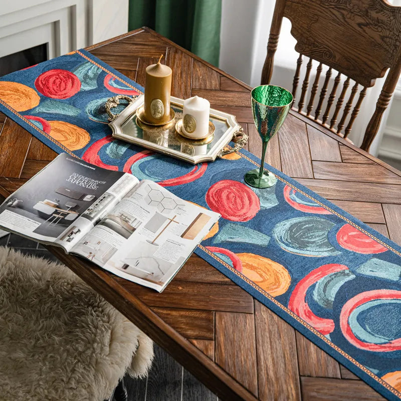 Sur une table en bois, on peut voir un chemin de table orné de motifs floraux colorés, accompagné de chandelles et d'un verre décoratif, ainsi qu'un magazine ouvert. Cette scène crée une ambiance chaleureuse et élégante dans la pièce.
