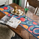 Sur une table en bois, on peut voir un chemin de table orné de motifs floraux colorés, accompagné de chandelles et d'un verre décoratif, ainsi qu'un magazine ouvert. Cette scène crée une ambiance chaleureuse et élégante dans la pièce.