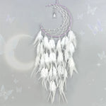 Cette image montre un capteur de rêves de lune blanche et argentée décoré de plumes blanches et violettes, suspendu sur un fond gris clair scintillant avec des papillons.