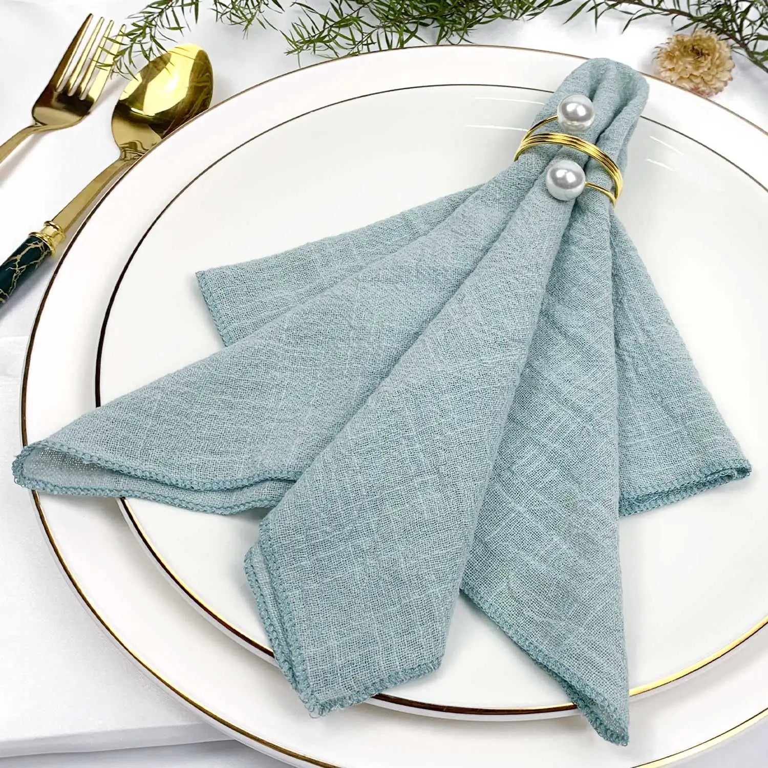 Serviette grise délicatement nouée avec des détails dorés, posée élégamment sur une assiette blanche avec des couverts dorés, le tout accompagné d'éléments végétaux.