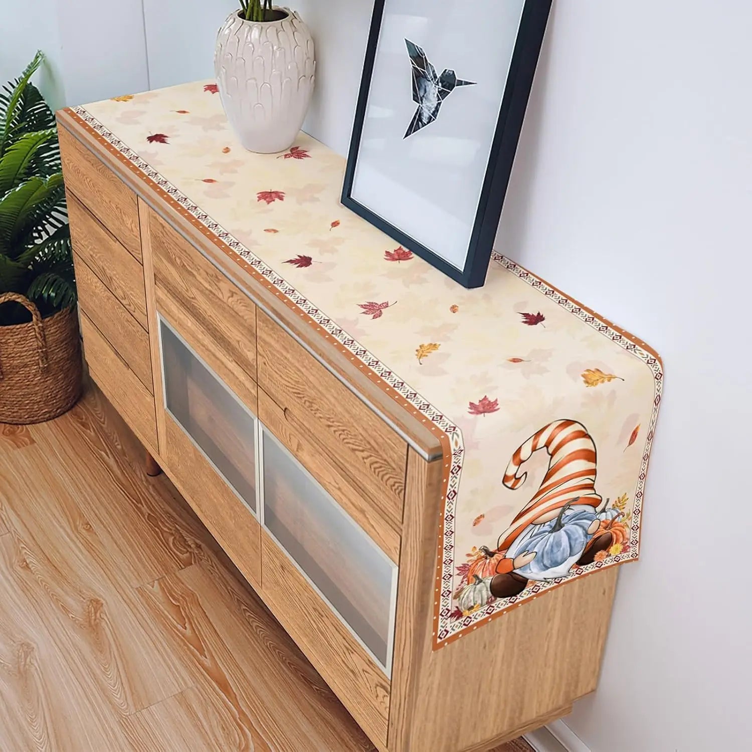 Un meuble en bois décoré d'une nappe aux motifs d'automne, avec une vase et un cadre mural, dans un décor de style scandinave.