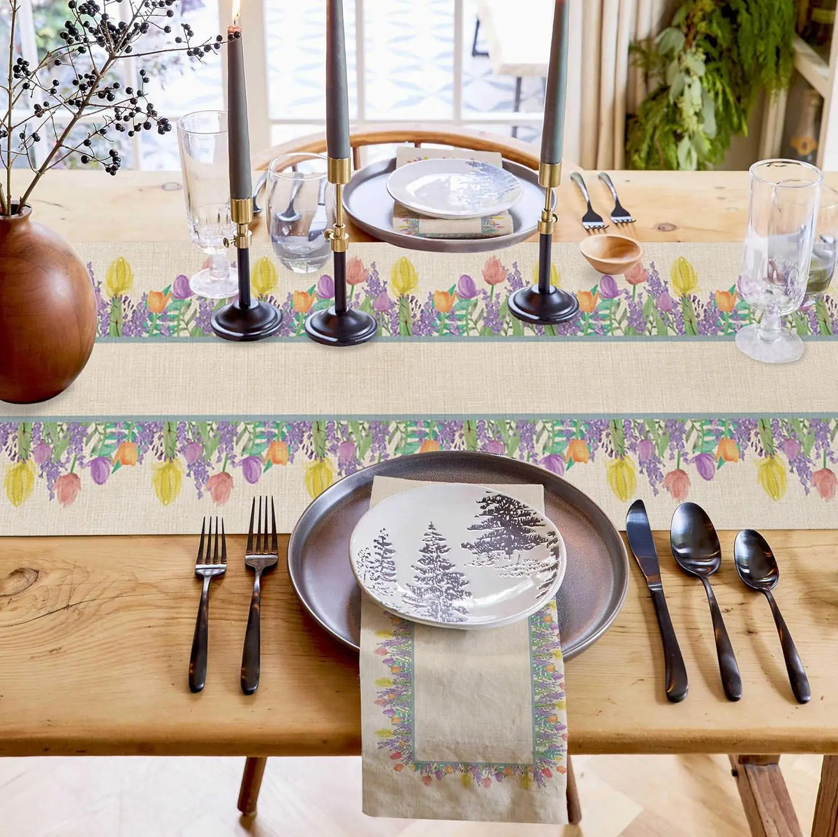 Une table joliment dressée avec des bougies, des verres, une nappe florale et une assiette avec un motif hivernal.