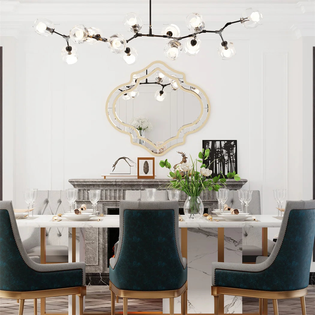 Salle à manger élégante avec un grand lustre moderne à plusieurs branches, un miroir décoratif encadré sur le mur, une table en marbre avec des chaises rembourrées en velours bleu, des fleurs dans un vase, et des décorations murales, créant une atmosphère chaleureuse et sophistiquée.