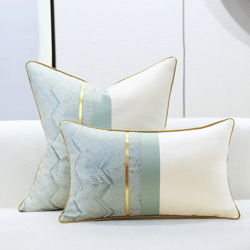 Deux oreillers décoratifs carrés de couleurs claires, avec des motifs graphiques et des bordures dorées, posés sur un lit blanc.