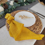 Table de cuisine décorée avec une nappe en lin jaune, un plateau en fibres tressées, des couverts et un plat en céramique blanche orné d'un élément doré.