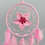 Un attrapeur de rêves rose en forme de fleur ornée de perles et de franges, suspendu dans un cadre circulaire.