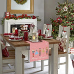 Cette image présente une table de salle à manger joliment décorée pour Noël, avec des ornements rouges et verts, des bougies, et un arbre de Noël dans un coin. La table est recouverte d'un chemin de table avec des motifs de personnages de Noël et l'inscription "Joyeux Noël".