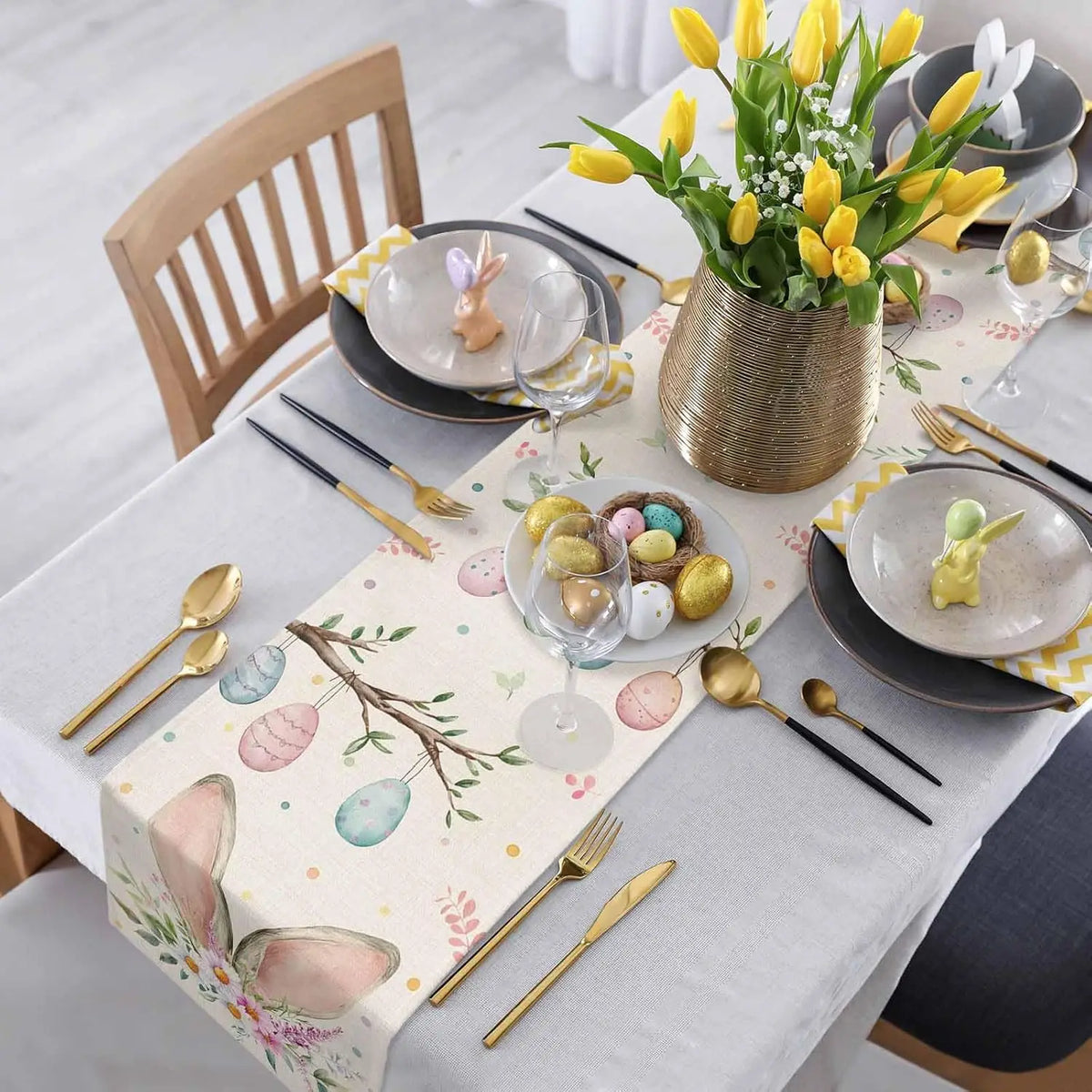 Cette image représente une belle table de Pâques avec des décors floraux printaniers, des couverts dorés et une assiette avec des œufs de Pâques colorés.