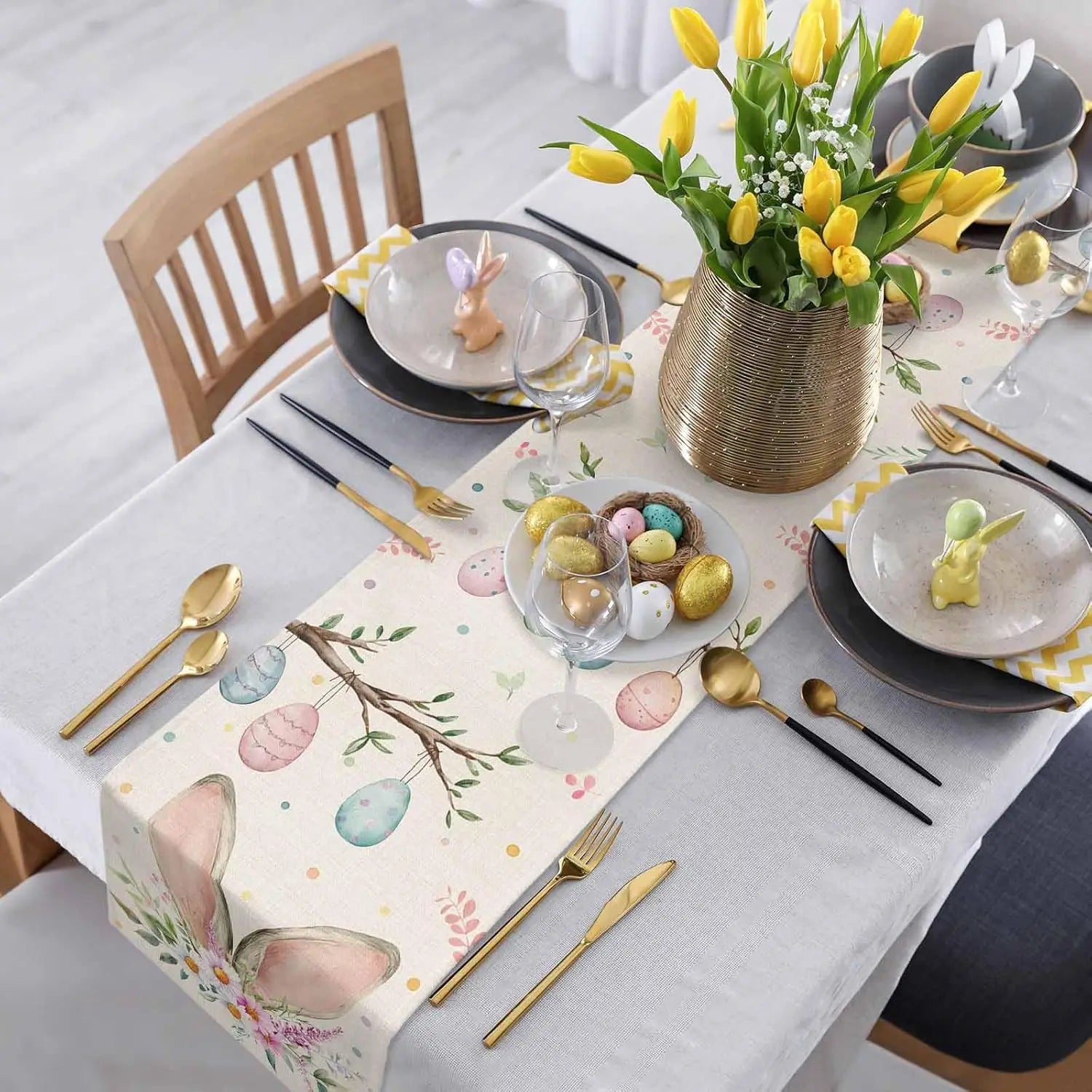 Cette image représente une belle table de Pâques avec des décors floraux printaniers, des couverts dorés et une assiette avec des œufs de Pâques colorés.