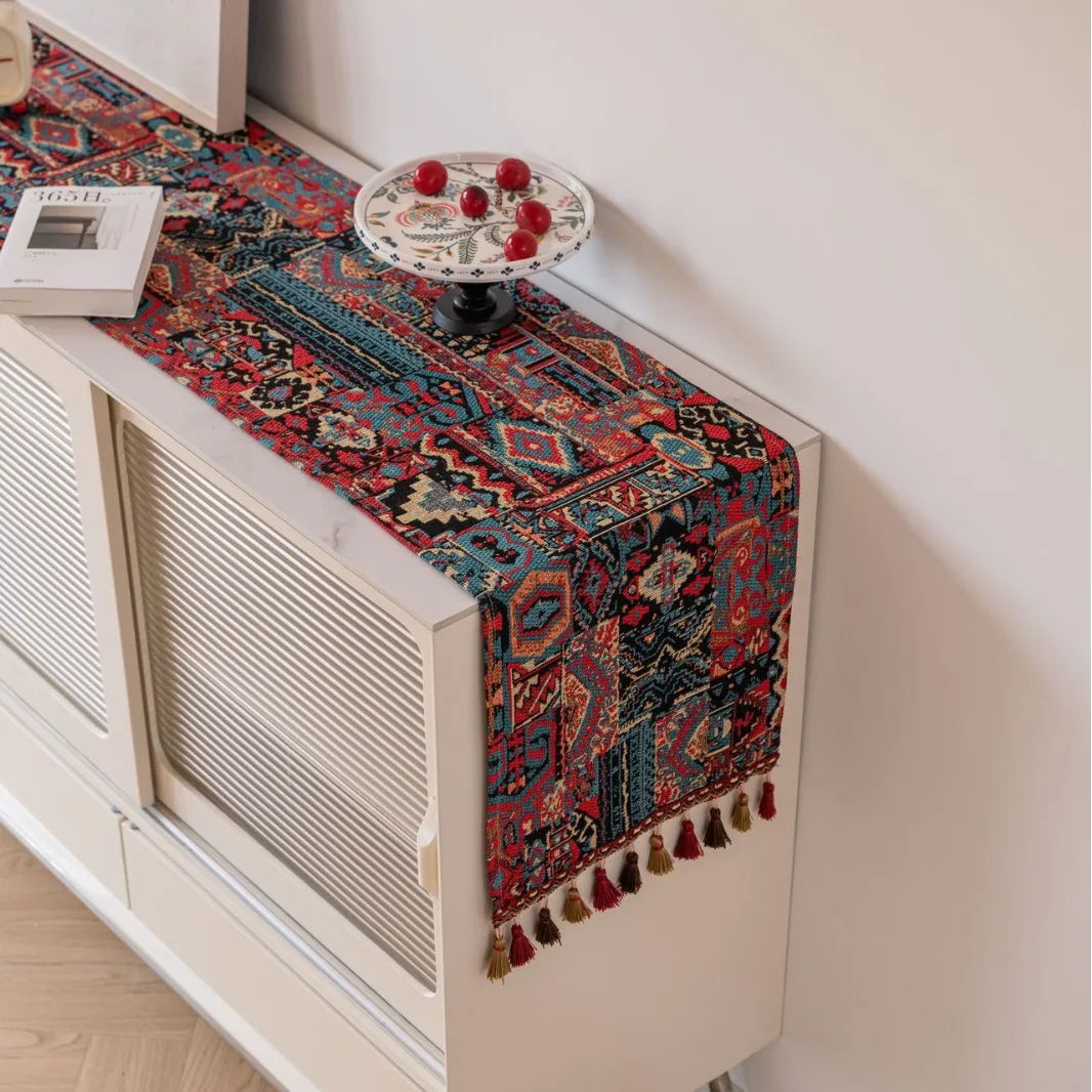 Un tapis de table coloré et décoratif avec des motifs ornementaux et des pompons franges, posé sur un meuble blanc, complétant l'ambiance chaleureuse et éclectique de la pièce.