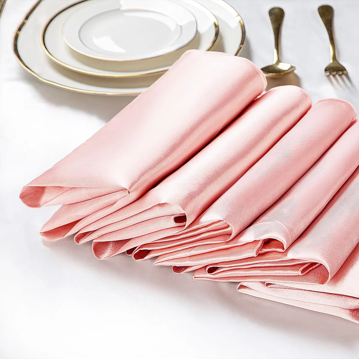 Une pile de serviettes de table en satin rose, disposées avec élégance auprès d'assiettes et de couverts dorés, créant une mise en place raffinée.