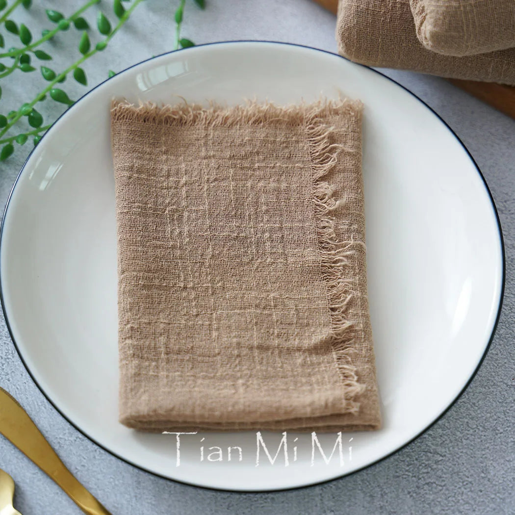 Une serviette de table en lin naturel avec des franges sur une assiette blanche, entourée de feuillage vert et d'accessoires dorés.