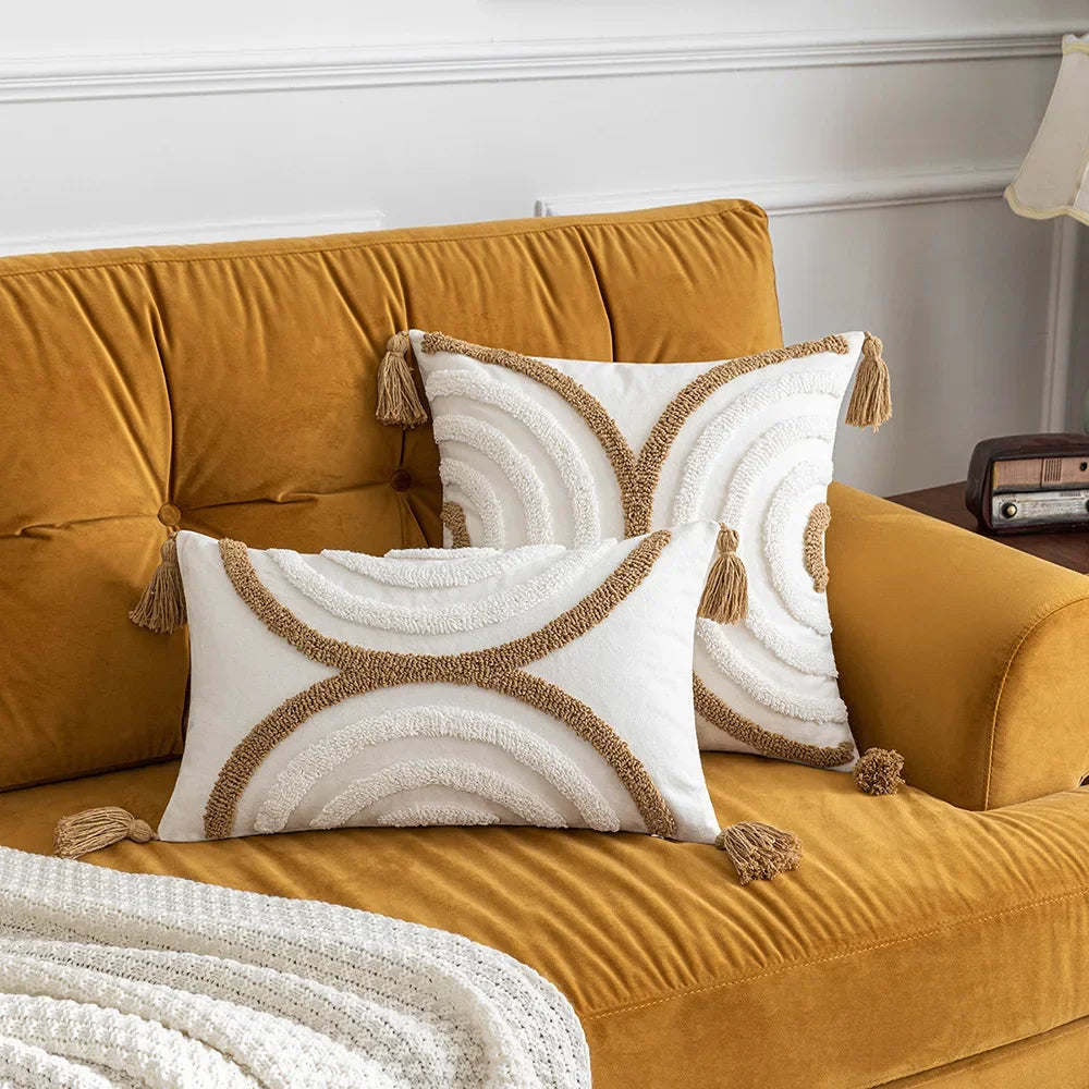 Deux coussins décoratifs blancs et beiges ornés de franges et de motifs graphiques sur un canapé jaune ocre dans un salon moderne.
