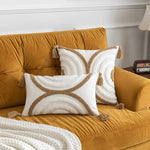 Deux coussins décoratifs blancs et beiges ornés de franges et de motifs graphiques sur un canapé jaune ocre dans un salon moderne.