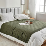 Une couverture douce et confortable en couleur verte, avec un motif de texture tressée, reposant sur un lit blanc dans une chambre lumineuse et accueillante.