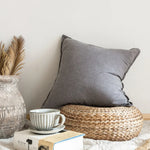 Un coussin gris confortable sur un tabouret en rotin, avec une tasse et un livre sur une surface en bois.