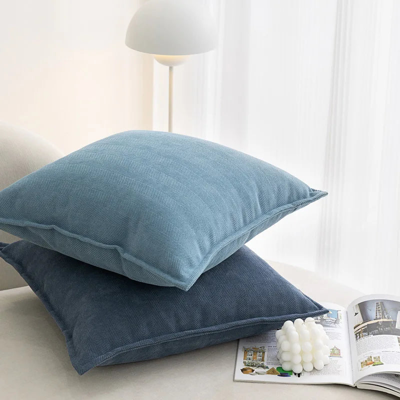 Deux oreillers décoratifs bleu-gris en textile avec une texture en chevrons sur une table de chevet à côté d'une lampe de lecture et d'un magazine ouvert.