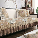 Cette image représente un canapé d'intérieur élégant et luxueux, avec des coussins et une couverture ornés de motifs floraux dorés sur un fond beige. Le canapé est placé dans un intérieur moderne avec des éléments décoratifs en bois sombre.