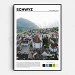 L'image montre une vue aérienne d'une ville suisse médiévale avec ses toits rouges, ses clochers d'églises et ses montagnes à l'arrière-plan. C'est la ville de Schwyz, connue pour son architecture historique et sa position dans une vallée entourée de montagnes.