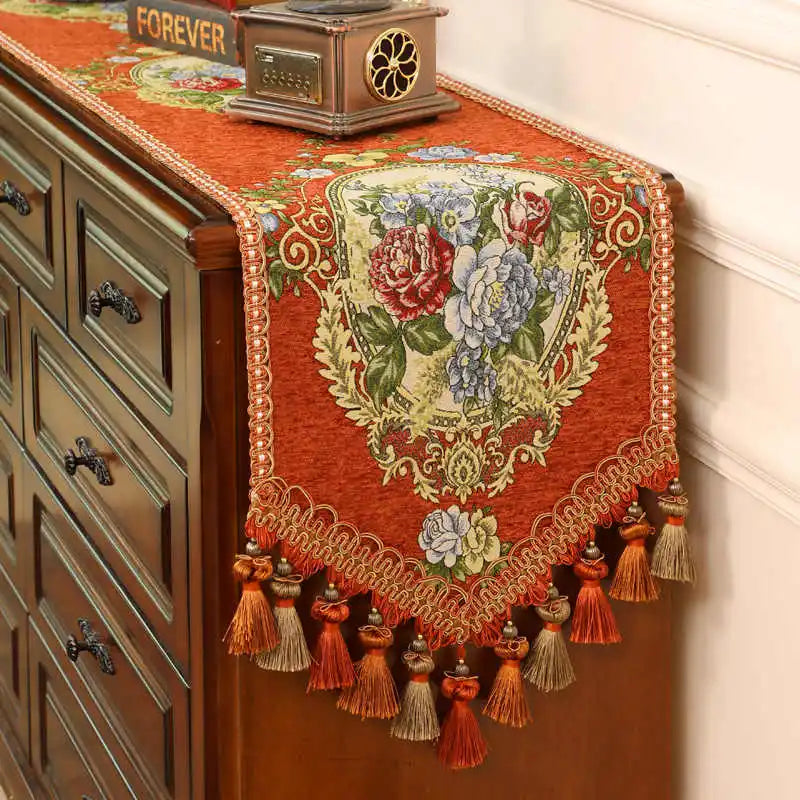 Un magnifique chemin de table brodé de fleurs colorées et de glands décoratifs sur un meuble ancien ajoute une touche élégante et vintage à cette scène.