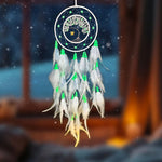 Un attrape-rêves lumineux avec un arbre de vie en son centre, décoré de plumes blanches et vertes, suspendu dans un ciel nocturne enneigé.