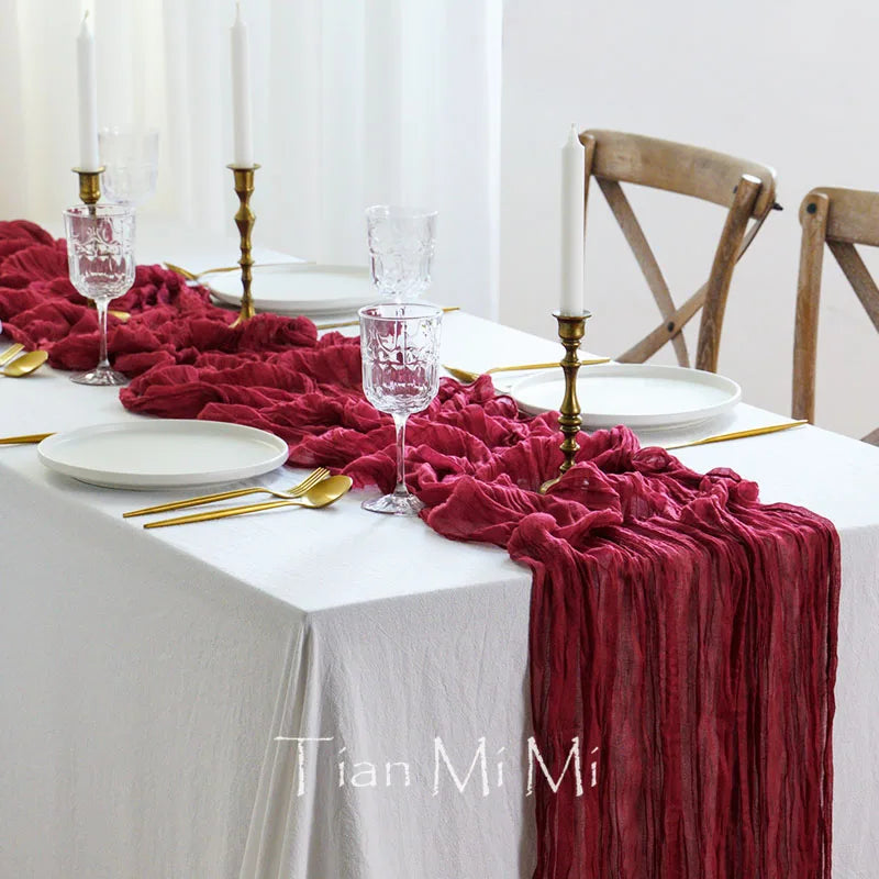 Table élégamment dressée avec une nappe blanche, des couverts dorés, des bougies et des verres de vin. Au centre, un chemin de table en velours rouge foncé apporte une touche chaleureuse et luxueuse à la décoration.