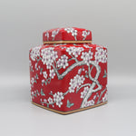 Un pot décoratif en céramique rouge vif orné de délicates fleurs de cerisier blanches, dégageant une atmosphère traditionnelle et élégante.