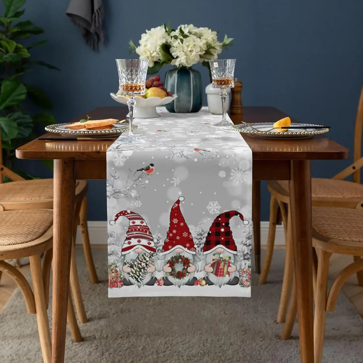 Une table festive avec une nappe de Noël ornée de gnomes mignons, de boules, de guirlandes et d'un bouquet de fleurs blanches au centre.