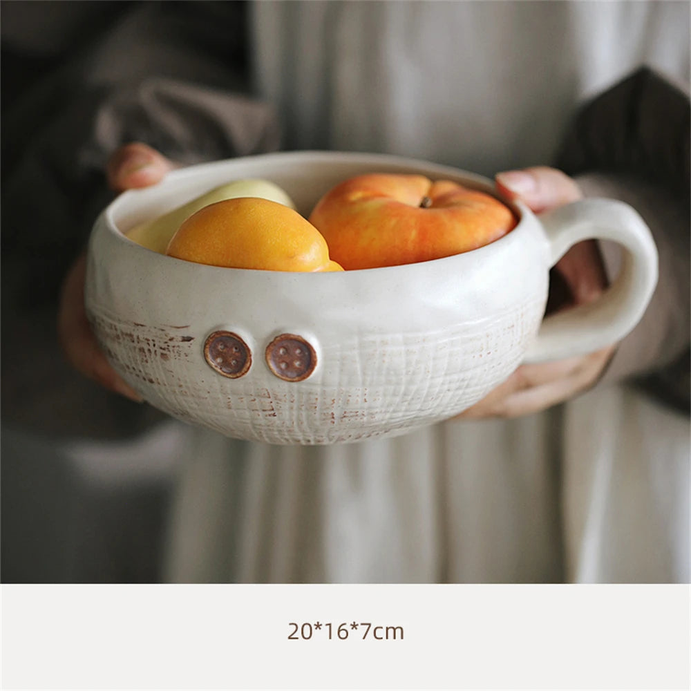 Bol en céramique blanche avec des accents de boutons en argile contenant différents fruits oranges, y compris une orange et une pomme.