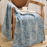 Élégante couverture en coton bleu orné de motifs paisley, posée sur un fauteuil vintage en bois, accompagnée d'une tasse de café sur une pile de disques vinyl.