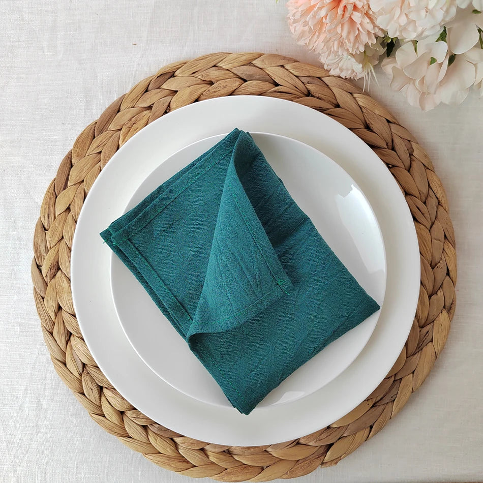 Une serviette verte foncée pliée sur une assiette blanche posée sur un dessous de plat en fibres tressées, avec des fleurs d'hortensias roses en arrière-plan.