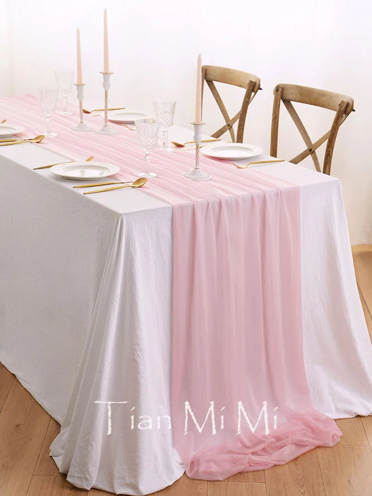 La table de salle à manger est élégamment dressée avec une nappe blanche et rose, des chandeliers et des couverts dorés, reflétant une ambiance raffinée et délicate.