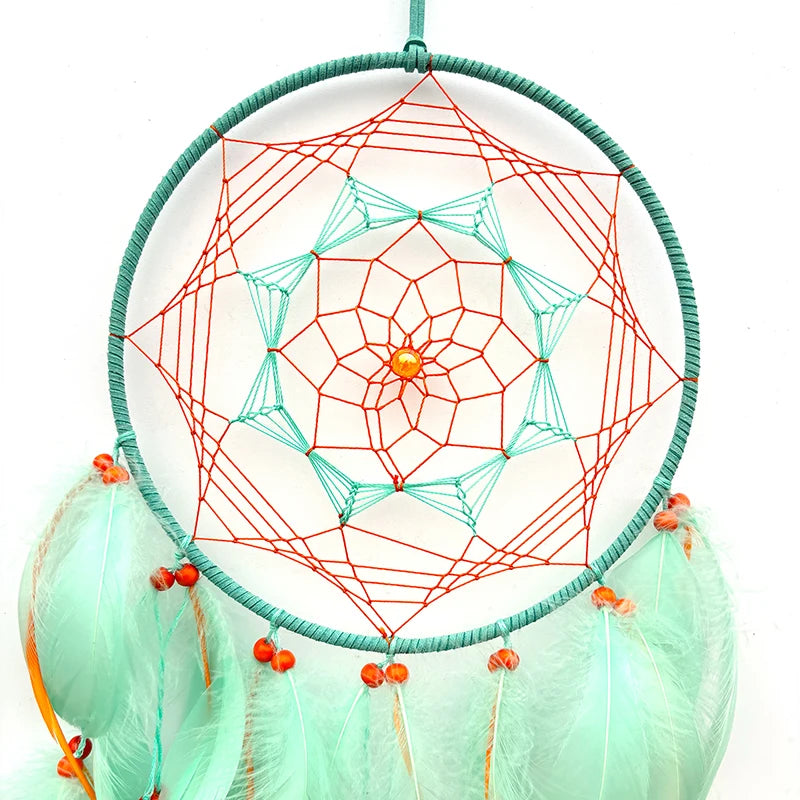 Un magnifique capteur de rêves avec un motif géométrique en spirale coloré, composé de fils rouges et vert menthe, surmonté d'une pierre orange et entouré de jolies plumes teintées.