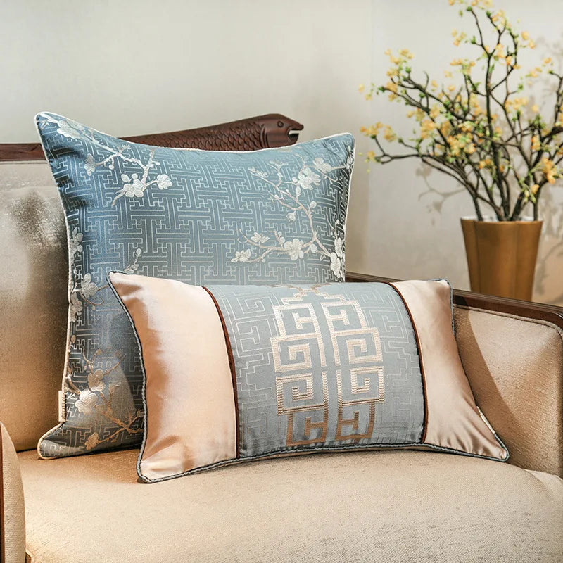 Deux coussins décoratifs de style asiatique avec motifs géométriques et floraux, posés sur un canapé beige, avec une plante en fleurs à l'arrière-plan.