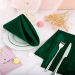 Des serviettes de table en satin vert foncé, accompagnées de couverts en métal et d'un livre de mémoire, créent une table de fête élégante avec des touches de couleurs pastel.