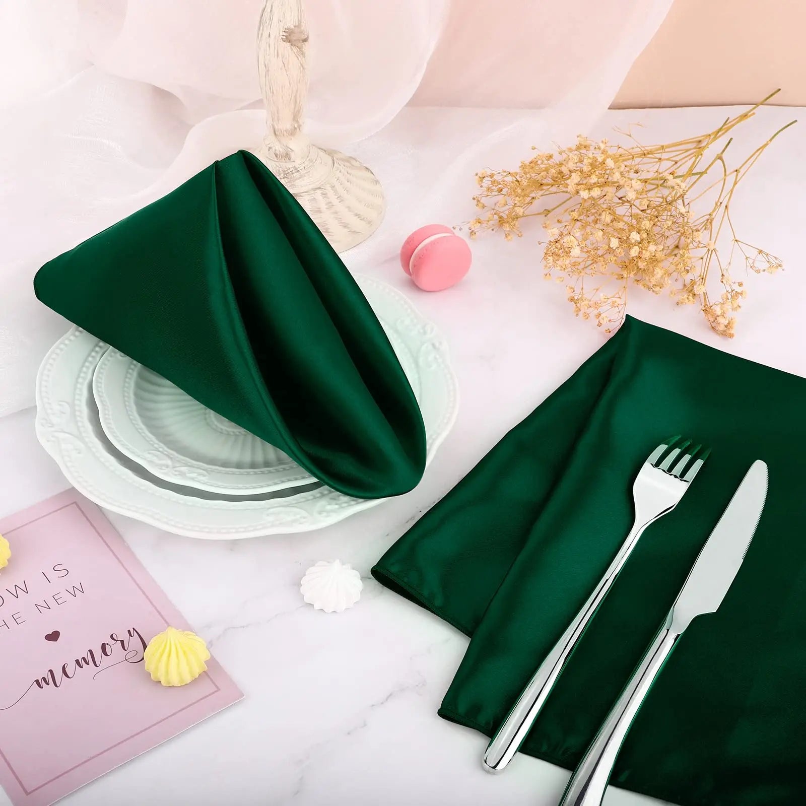 Des serviettes de table en satin vert foncé, accompagnées de couverts en métal et d'un livre de mémoire, créent une table de fête élégante avec des touches de couleurs pastel.