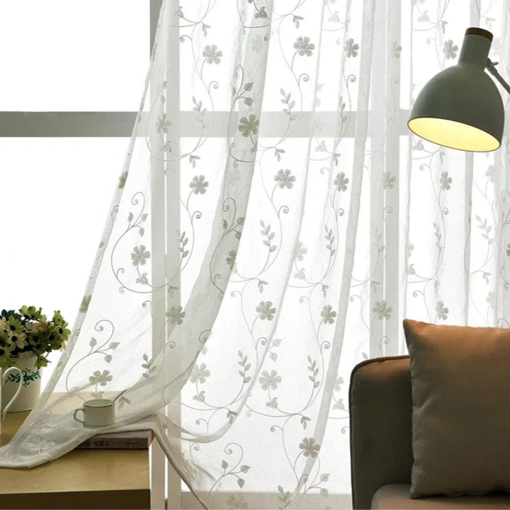 Image d'un salon bien éclairé avec des rideaux blancs en textile de haute qualité, couvrant une grande fenêtre pour un traitement optique optimal. Le design intérieur comprend un canapé-lit confortable, une lampe moderne et des plantes d'intérieur luxuriantes, reflétant un style de vie serein et élégant.