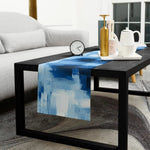 Jolie table basse en bois noir avec un chemin de table bleu abstraite posé dessus, accompagnée d'une horloge et d'un service à thé sur un tapis gris dans un salon confortable.