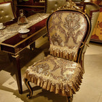 Chaise en bois de style vintage avec un dossier orné de motifs floraux et de franges dorées, placée dans un environnement meublé avec soin, reflétant une atmosphère élégante et chaleureuse.
