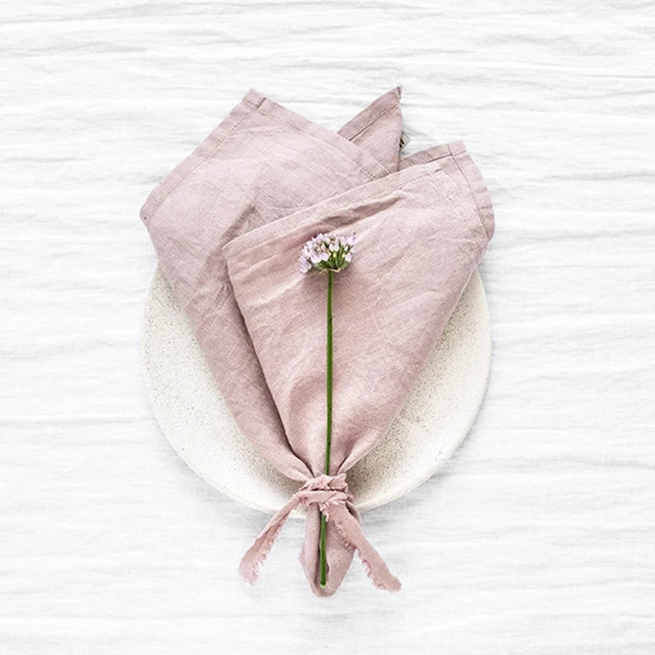 Une serviette de lin rose pâle repose sur un cercle blanc, avec une petite fleur blanche nouée au centre.