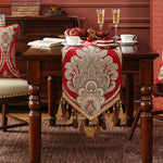 Une nappe colorée rouge et dorée, ornée de motifs élégants et de glands décoratifs, est posée sur une table en bois sombre, accompagnée d'assiettes, de verres et d'un gâteau, créant une ambiance chaleureuse et festive.