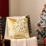 Un coussin décoratif de Noël en velours doré arborant un beau motif de flocon de neige trône sur un fauteuil, devant un sapin de Noël scintillant de guirlandes.