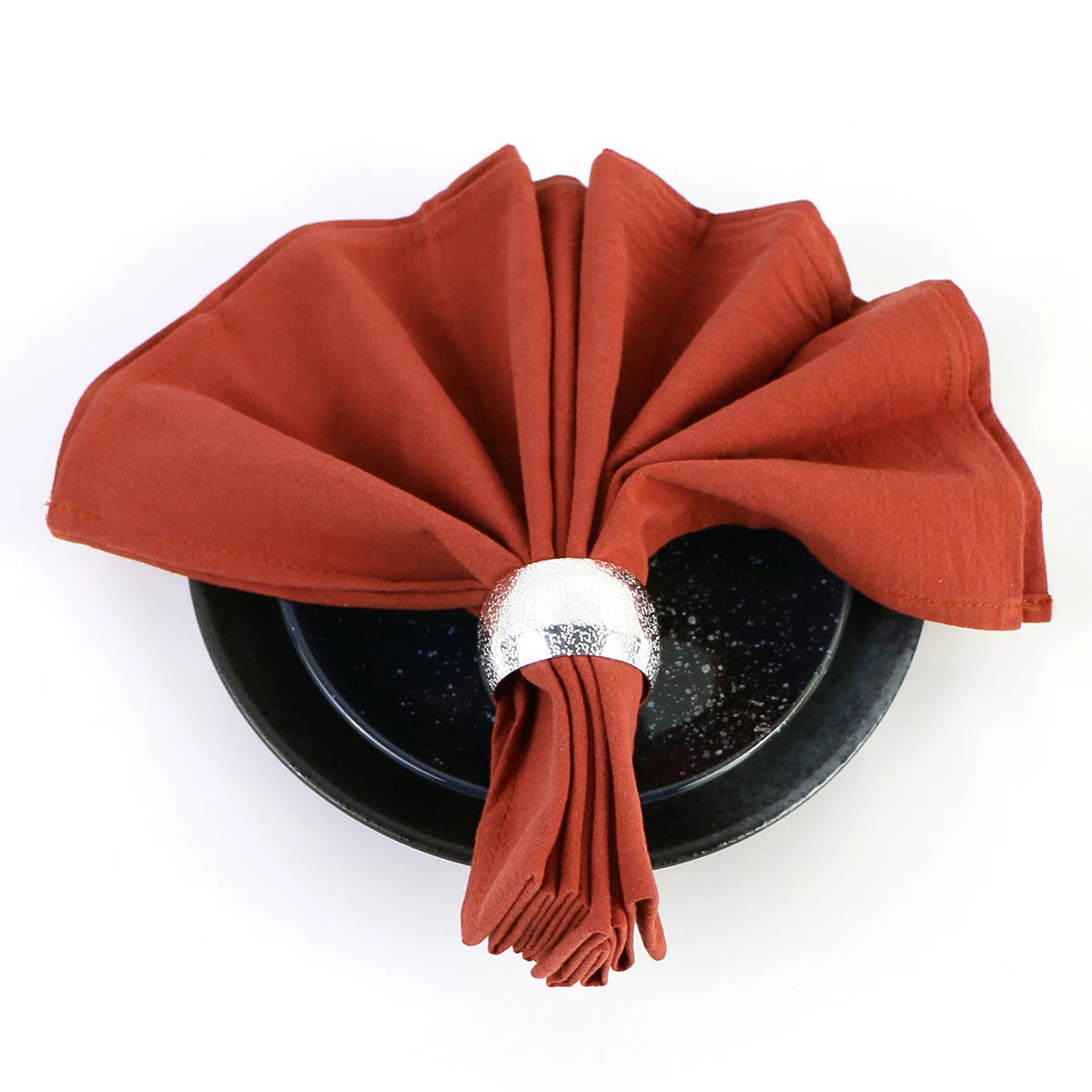 Serviette pliée en éventail de couleur rouge brique, posée sur un plat noir avec un anneau décoratif en métal argenté.