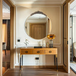 Un miroir rond doré orné de fleurs sculptées sur un meuble élégant en bois clair avec des tiroirs, placé dans un couloir lumineux avec un sol en bois.